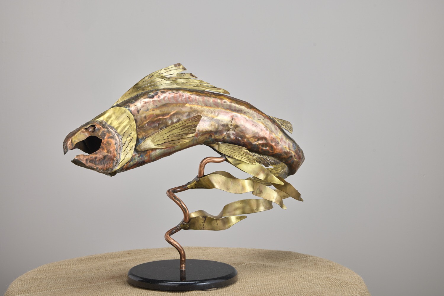Copper and brass fish sculpture leaping from a ribbon of metal on a round base